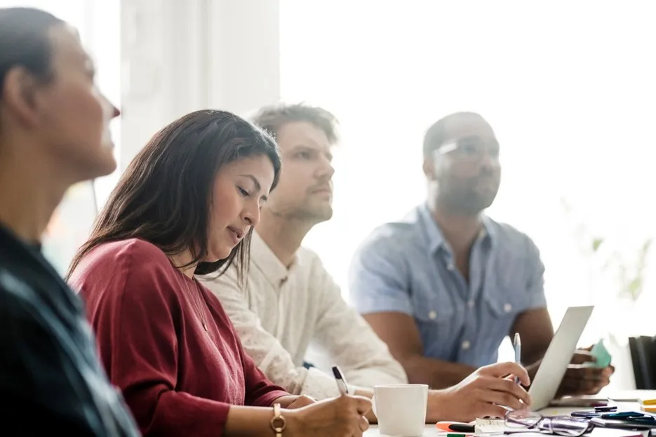A group of people in a meeting, one person writes while others listen.