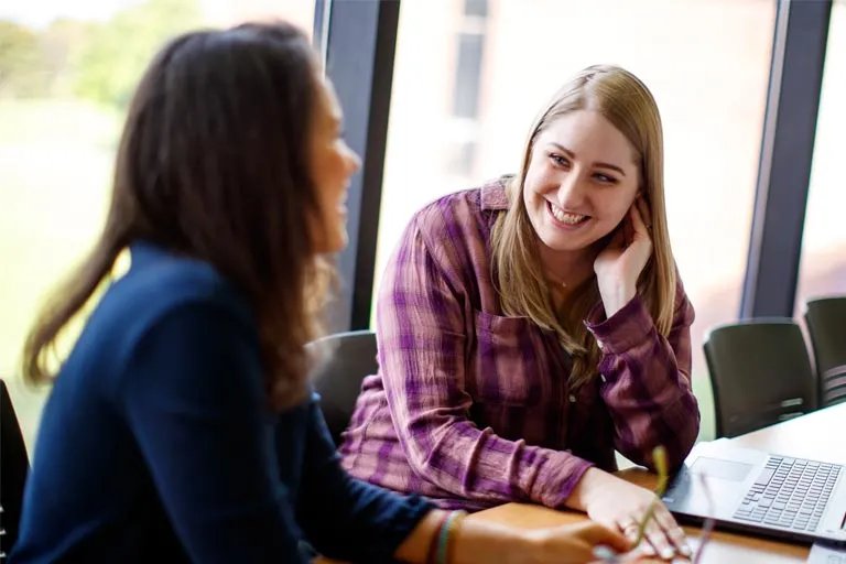 Two young women smiling, one in a plaid shirt, engaging in a discussion at a table.