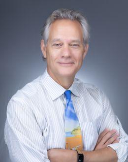 Smiling man with graying hair, striped shirt, and colorful tie, arms crossed.
