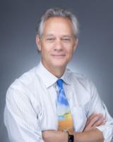 Smiling man with graying hair, striped shirt, and colorful tie, arms crossed.